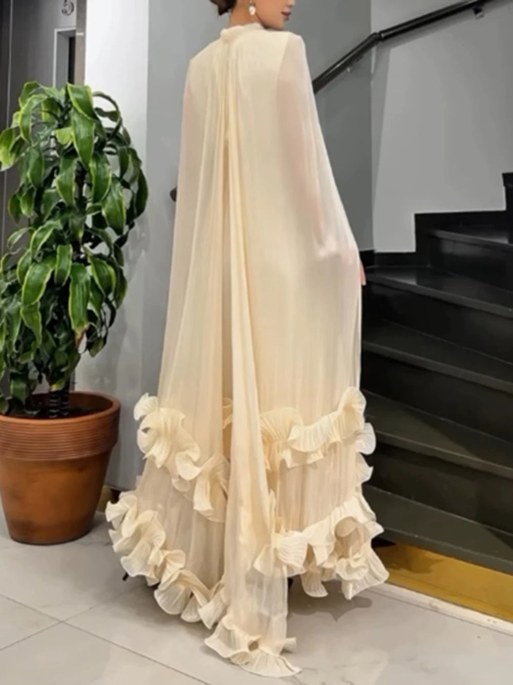 Woman wearing a long, flowing beige dress with ruffled hem in an indoor setting.