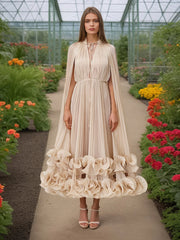 Woman in a beige dress with ruffled cape standing in a garden.
