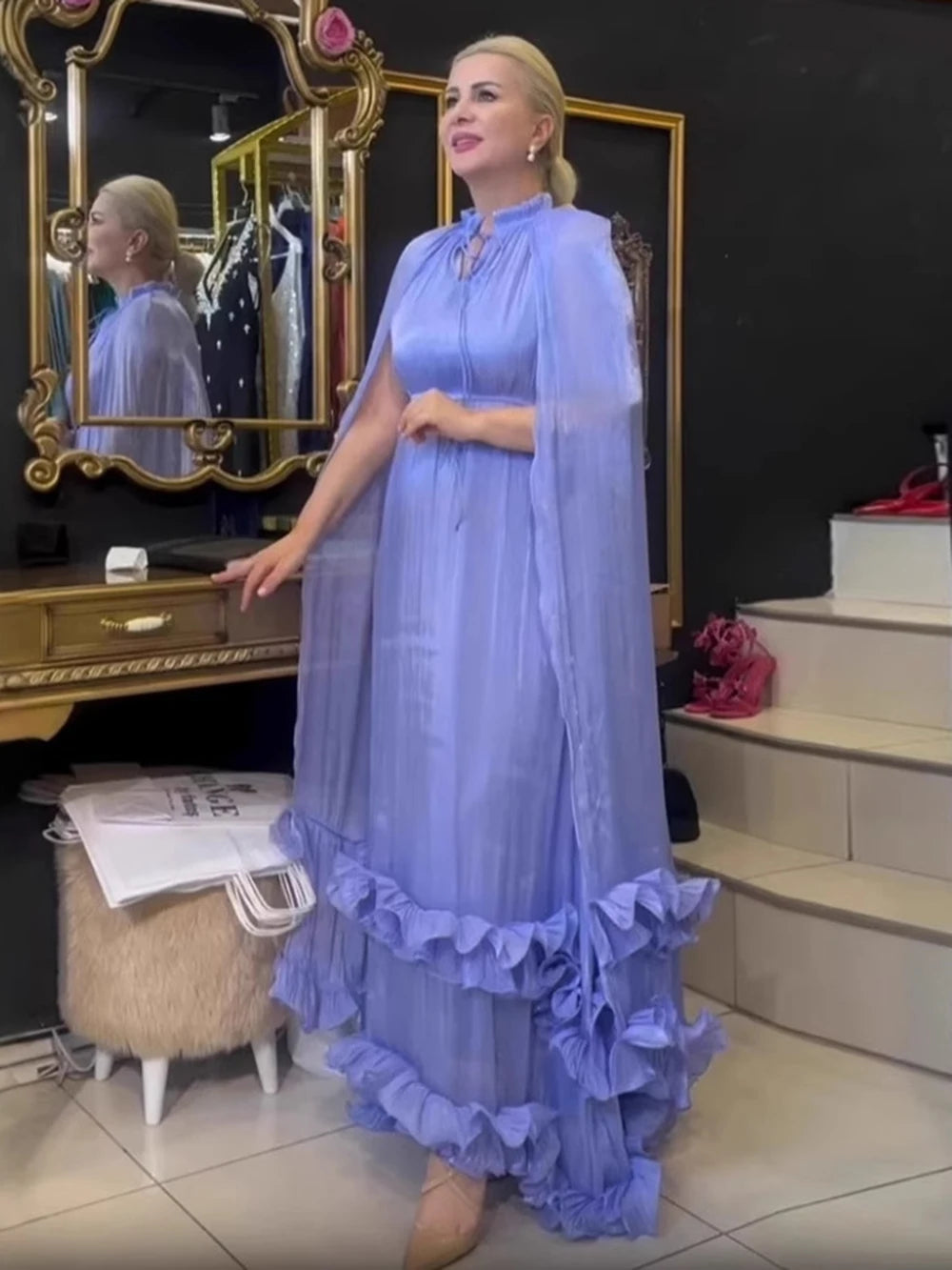 Woman in a light blue dress standing in a room with mirrors and furniture.
