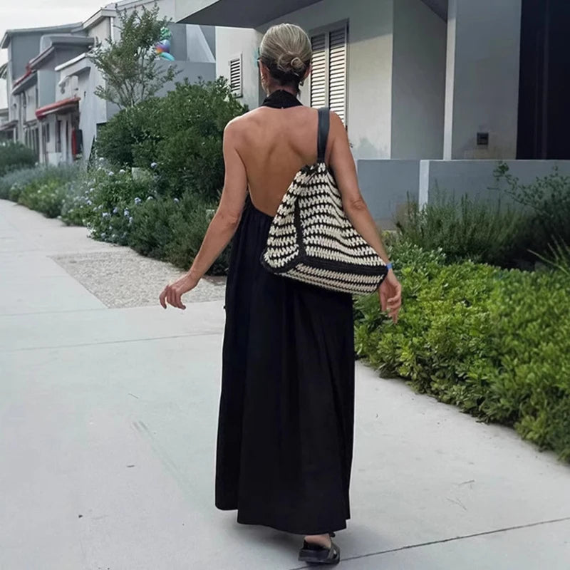 Woman in a black dress with an open back walking on a sidewalk, carrying a striped bag.
