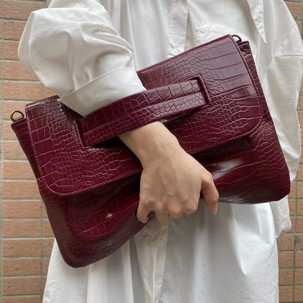 Person holding a maroon crocodile-patterned clutch bag against a brick wall.