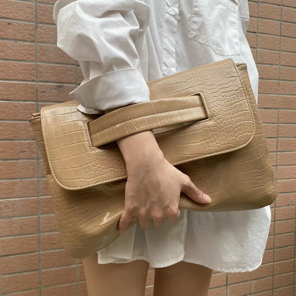 Beige handbag held by a person wearing a white shirt and shorts against a brick wall.