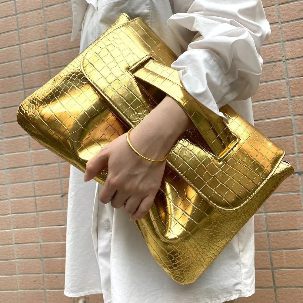 Gold clutch bag held by a person wearing a white outfit against a brick wall.