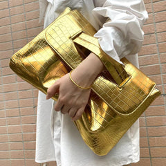Gold clutch bag held by a person wearing a white outfit against a brick wall.