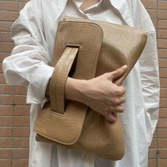 Person holding a beige crocodile-patterned clutch against a brick wall.