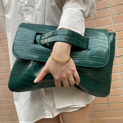 Person holding a green crocodile-patterned handbag against a brick wall.