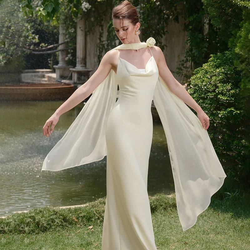 Woman in a flowing white dress standing outdoors near a pond with greenery.
