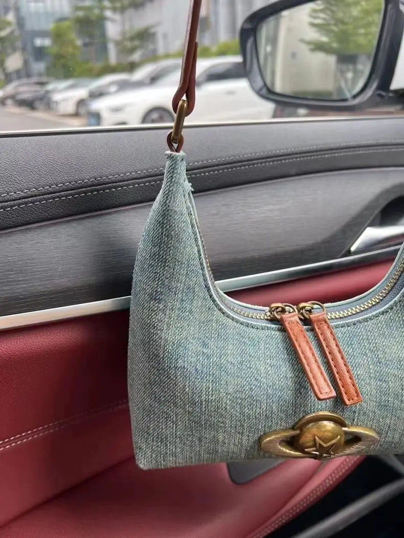 Denim handbag with leather straps hanging from a car mirror inside a vehicle.