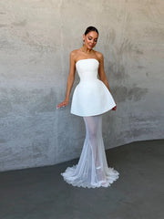 Woman wearing a strapless white dress with a fitted bodice and flowing skirt against a plain background.