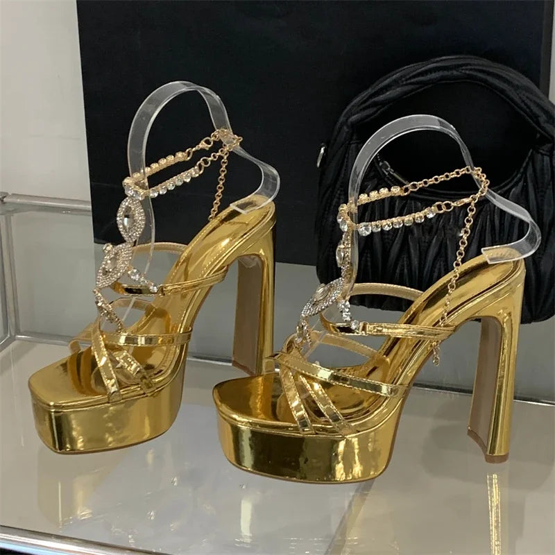Gold high-heeled shoes with clear straps and chain details on a reflective surface.