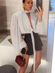 Woman taking a mirror selfie wearing a polka dot blouse and black shorts, holding a red handbag.