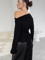 Person wearing a black off-shoulder top and black pants in a minimalistic setting.