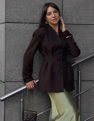 Woman in a dark brown blazer and light green skirt standing against a gray wall.