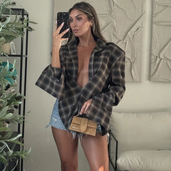 Woman taking a mirror selfie wearing a plaid shirt over a black top and denim shorts, holding a beige purse.