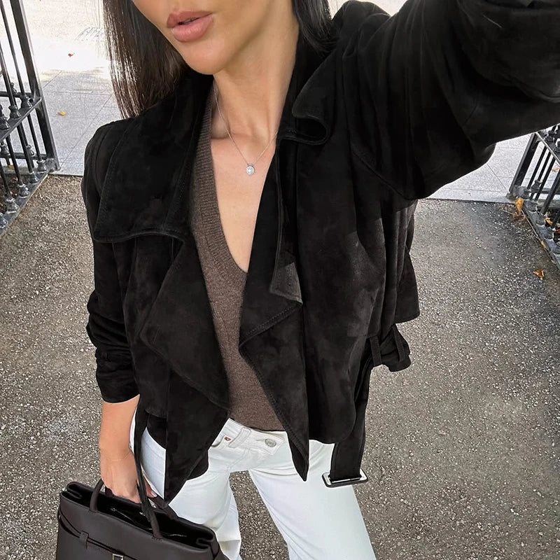 Person wearing a black suede jacket over a brown top and white pants, holding a black handbag.
