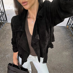Person wearing a black suede jacket over a brown top and white pants, holding a black handbag.