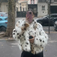 Person wearing a white fur coat with black and brown spots, taking a selfie on a street.