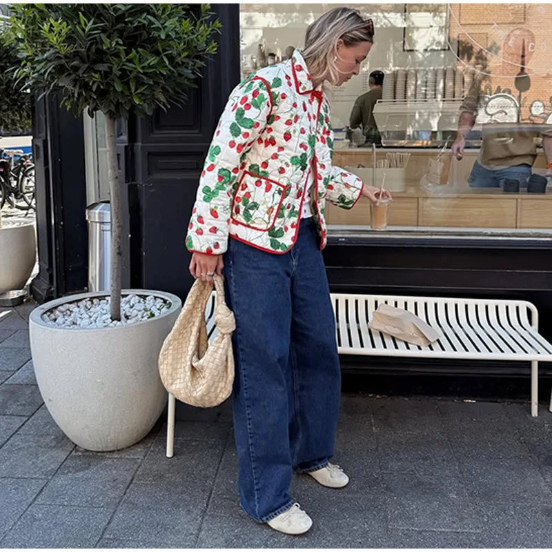 Person wearing a colorful jacket with cherry pattern, holding a drink, standing on a sidewalk.