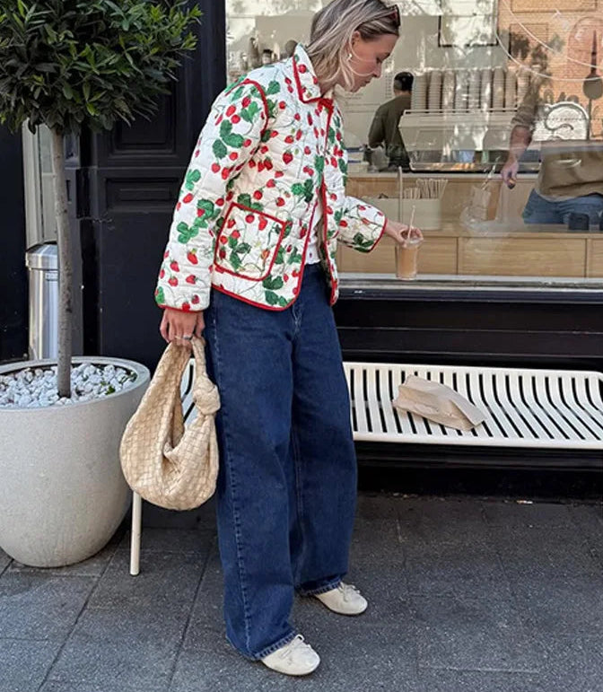 Person wearing a colorful jacket with cherry pattern, holding a drink, standing on a sidewalk.