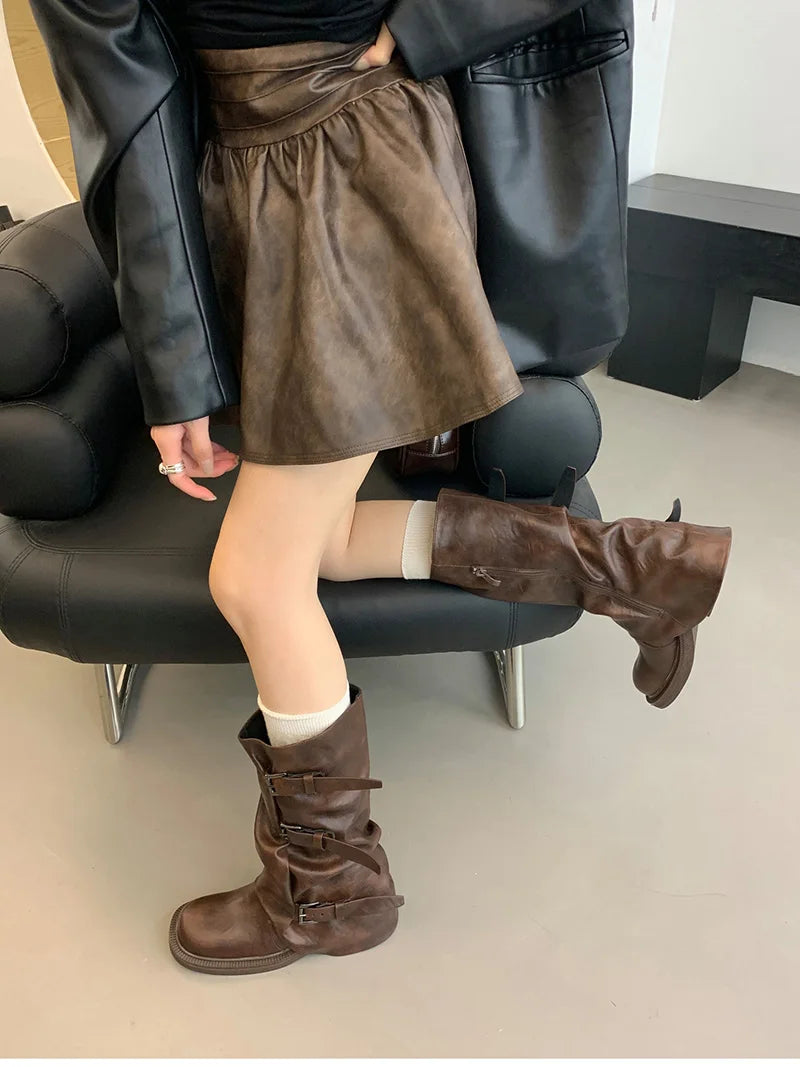 Person wearing a brown skirt and knee-high boots sitting on a black chair.