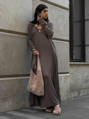 Woman in a long brown dress holding a beige bag against a gray building.