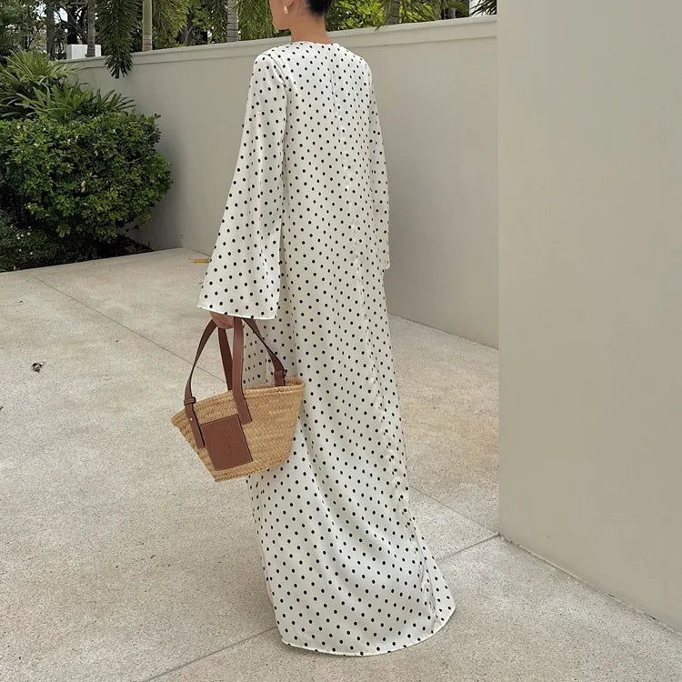 Person wearing a long white dress with black polka dots, holding a straw bag outdoors.