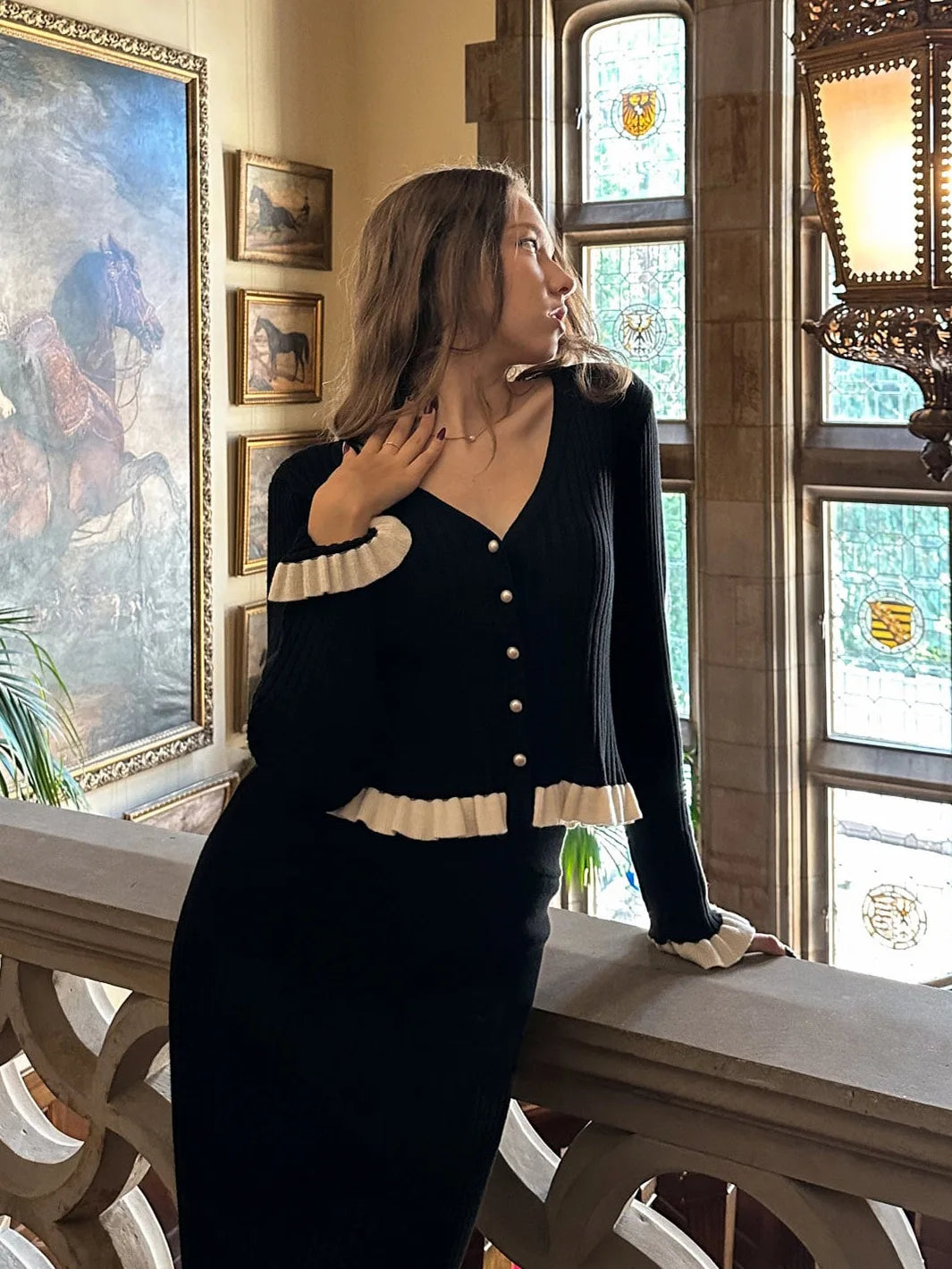 Woman in a black and white outfit standing in an ornate room with paintings and stained glass windows.
