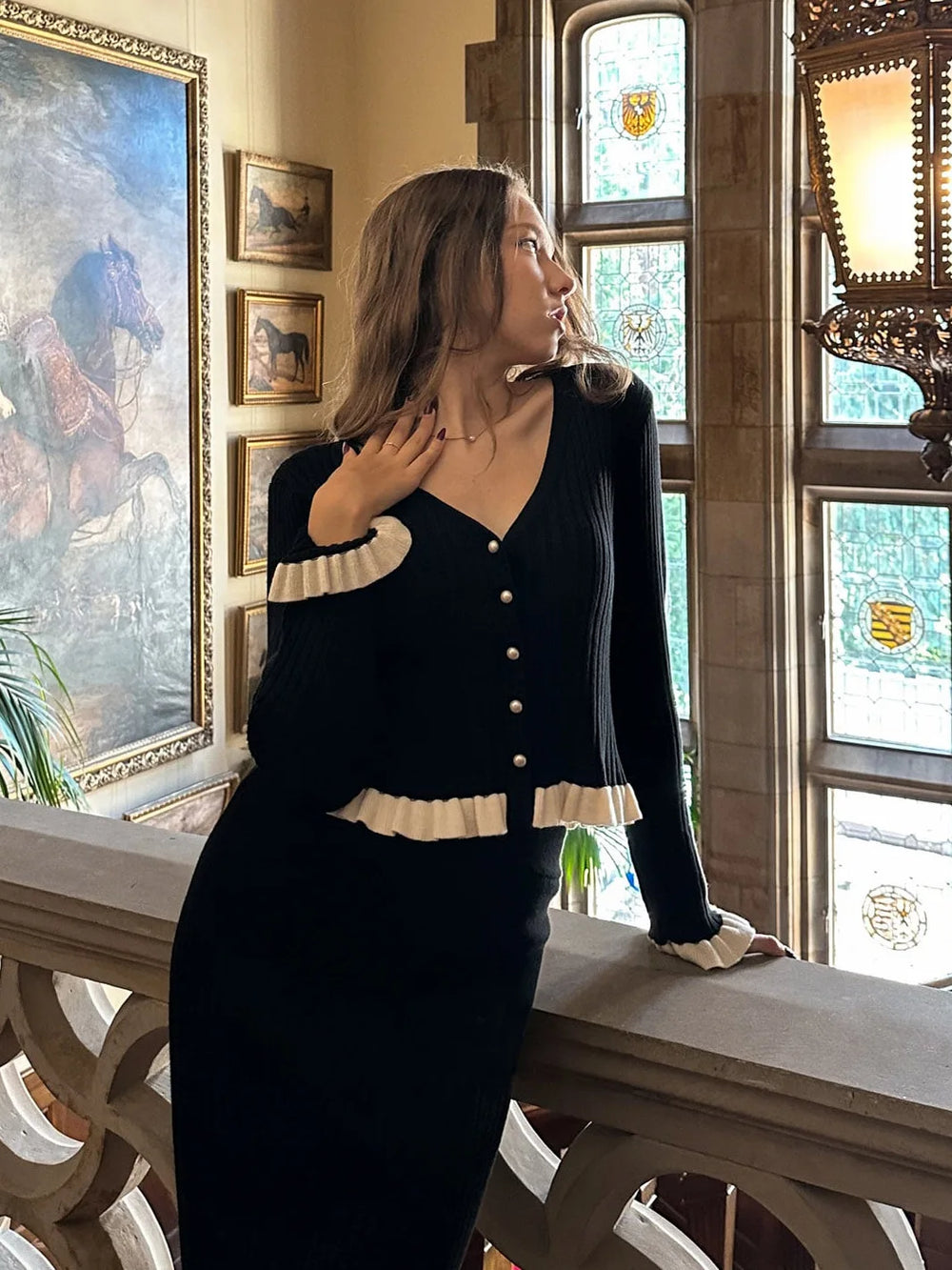 Woman in a black and white outfit standing in an ornate room with paintings and stained glass windows.