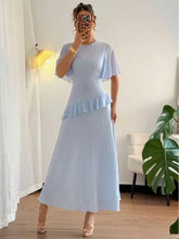 Woman wearing a light blue dress with ruffled sleeves and pleated skirt in a room with white curtains and a plant.