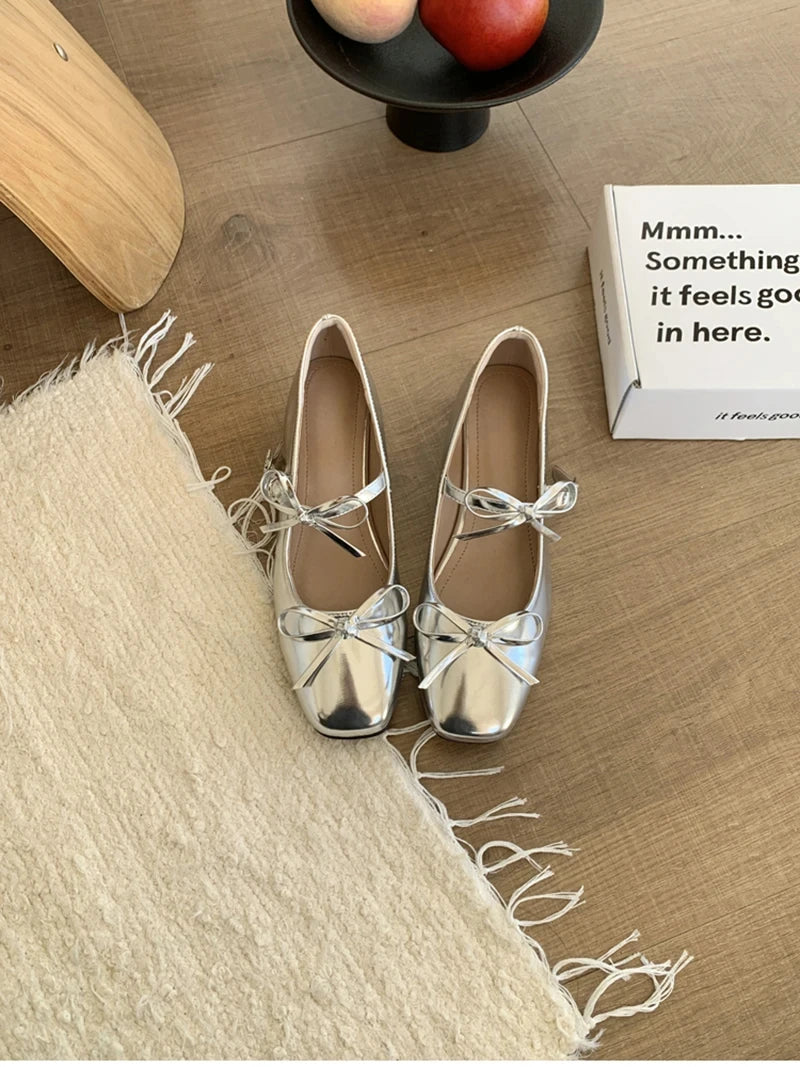 Silver ballet flats with bows on a wooden floor next to a box.