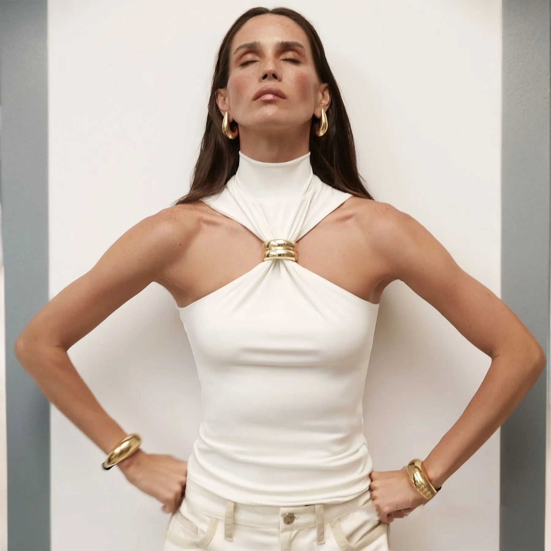 Woman wearing a white halter top with a gold accent against a plain background