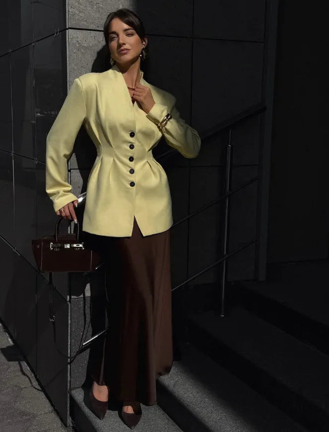 Woman in a light yellow blazer and brown pants standing against a dark wall.