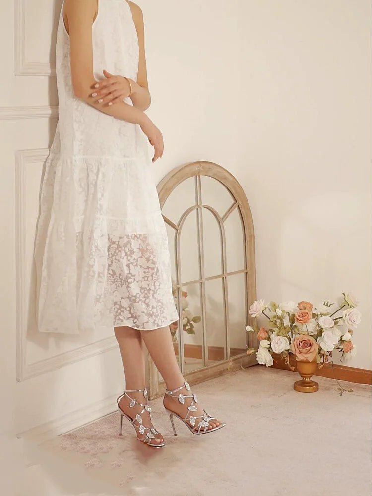 Woman in a white lace dress standing in a room with a mirror and flowers.
