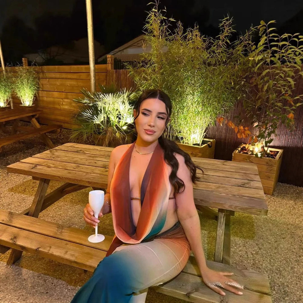 Woman sitting at a wooden picnic table outdoors with plants and lights in the background