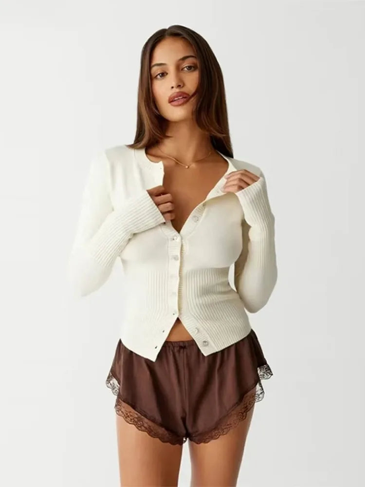 Woman wearing a white cardigan over a white top and brown shorts with lace details on a light gray background