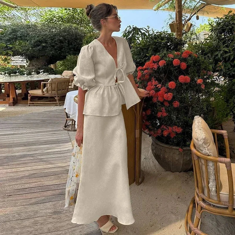 Woman in a white outfit standing in an outdoor setting with plants and furniture.