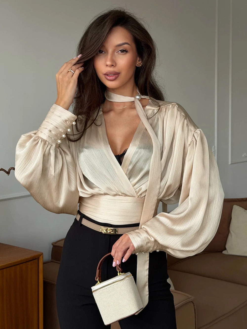 Woman in a stylish beige blouse holding a white handbag in a living room setting.