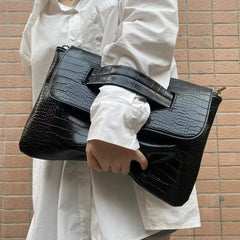 Person holding a black crocodile-patterned handbag against a brick wall.