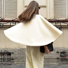 Person wearing a beige cape and holding a black bag on a street with stone walls and shutters.
