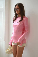 Woman wearing a pink long-sleeve top and white shorts against a white wall.