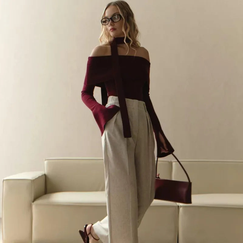Woman in a stylish outfit with a off-shoulder top and high-waisted pants, holding a handbag, standing against a neutral background.