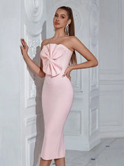 Woman wearing a pink strapless dress with a large bow detail against a white decorative wall.