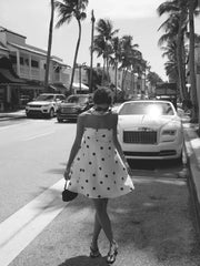 Woman in a polka dot dress walking on a street with palm trees and a luxury car in the background.
