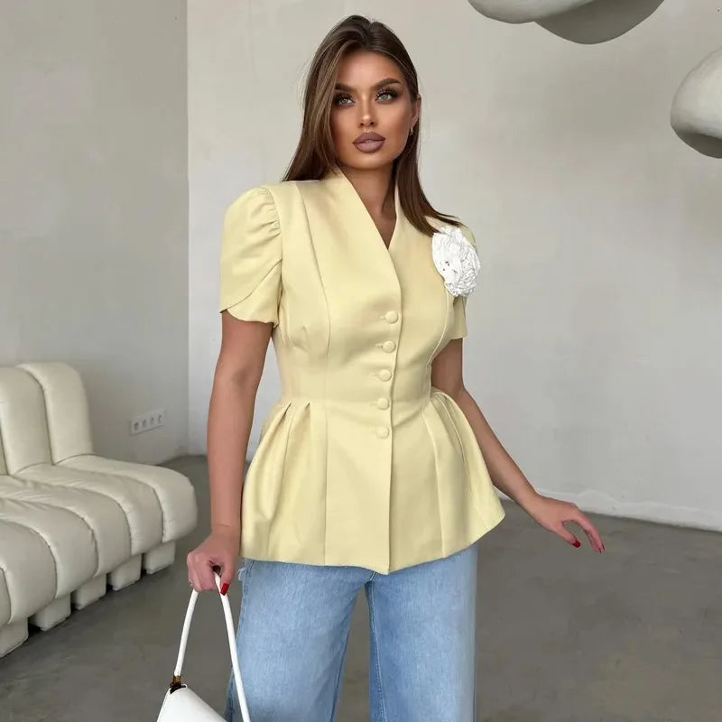 Woman wearing a yellow peplum top with a white flower detail, standing in a modern room.