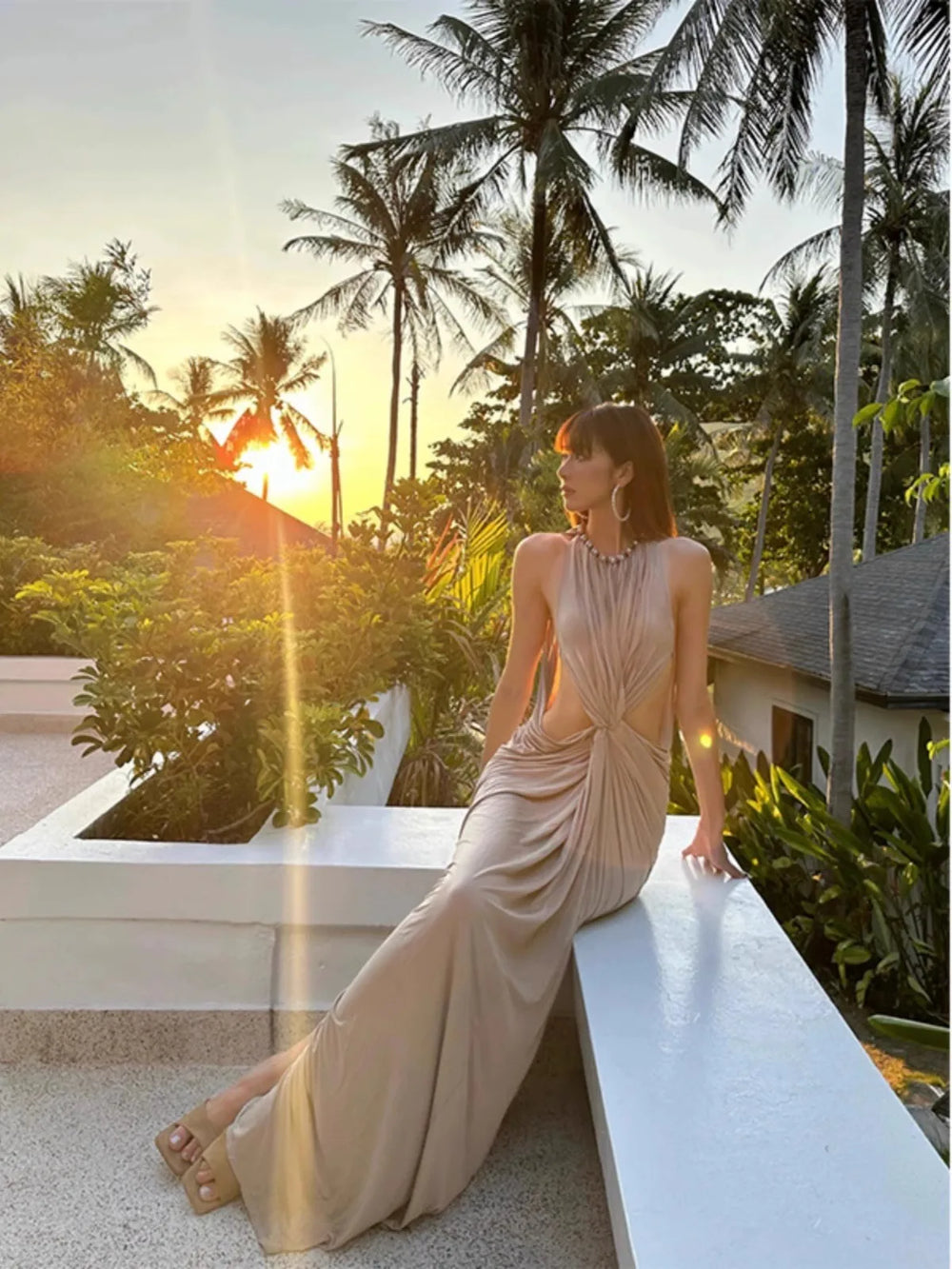 Woman in a beige dress sitting on a ledge with palm trees and sunset in the background