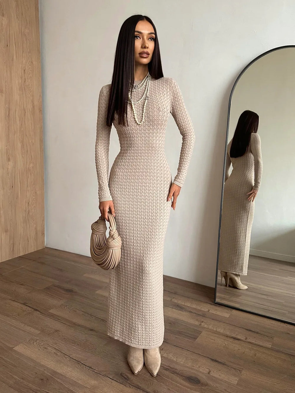 Woman in a long, light-colored knit dress standing in a room with wooden flooring and a mirror.