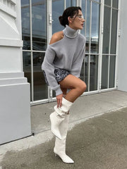 Person wearing a gray sweater, patterned shorts, and white knee-high boots standing in front of a glass door.
