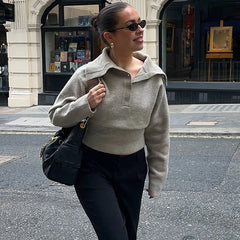 A woman wearing a knitted polo sweater, leaning casually near a storefront with tailored trousers and sunglasses