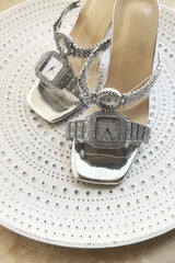 Silver high-heeled sandals with decorative straps on a white textured surface