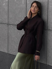 Woman wearing a black blazer and green pants against a gray tiled wall.